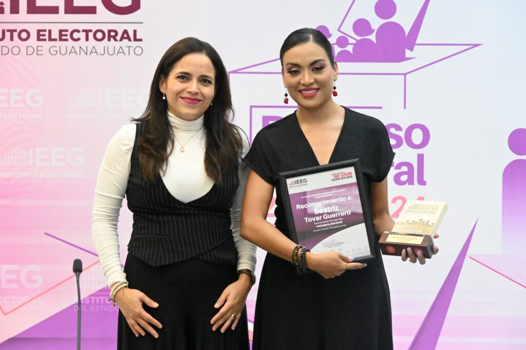 Two women stand together, one holding an award and a certificate. They are in front of a backdrop featuring the logo of an electoral institute.